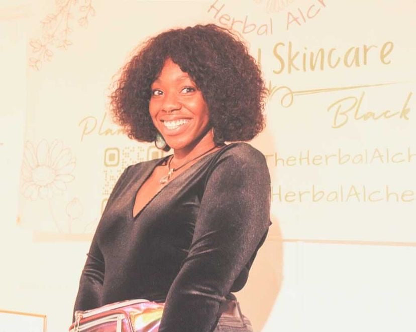 The Herbal Alchemist founder - Sade Smiling big in front of their banner which reads "The Herbal Alchemist" Natural Skincare Plant Powered. Black Owned
